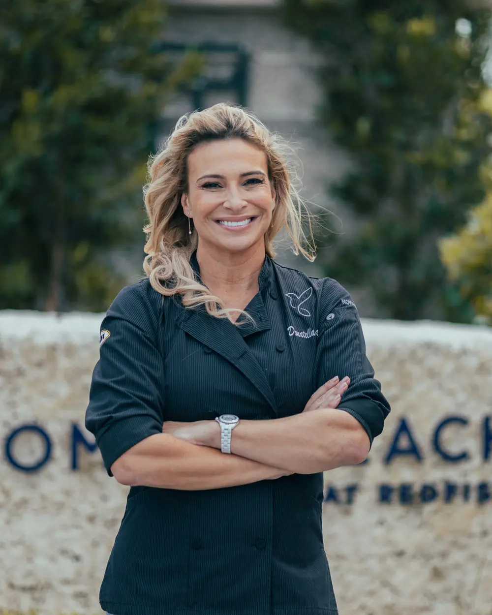 Donatella Arpaia in her chef’s jacket, arms crossed, smiling, the NOMA BEACH sign behind her