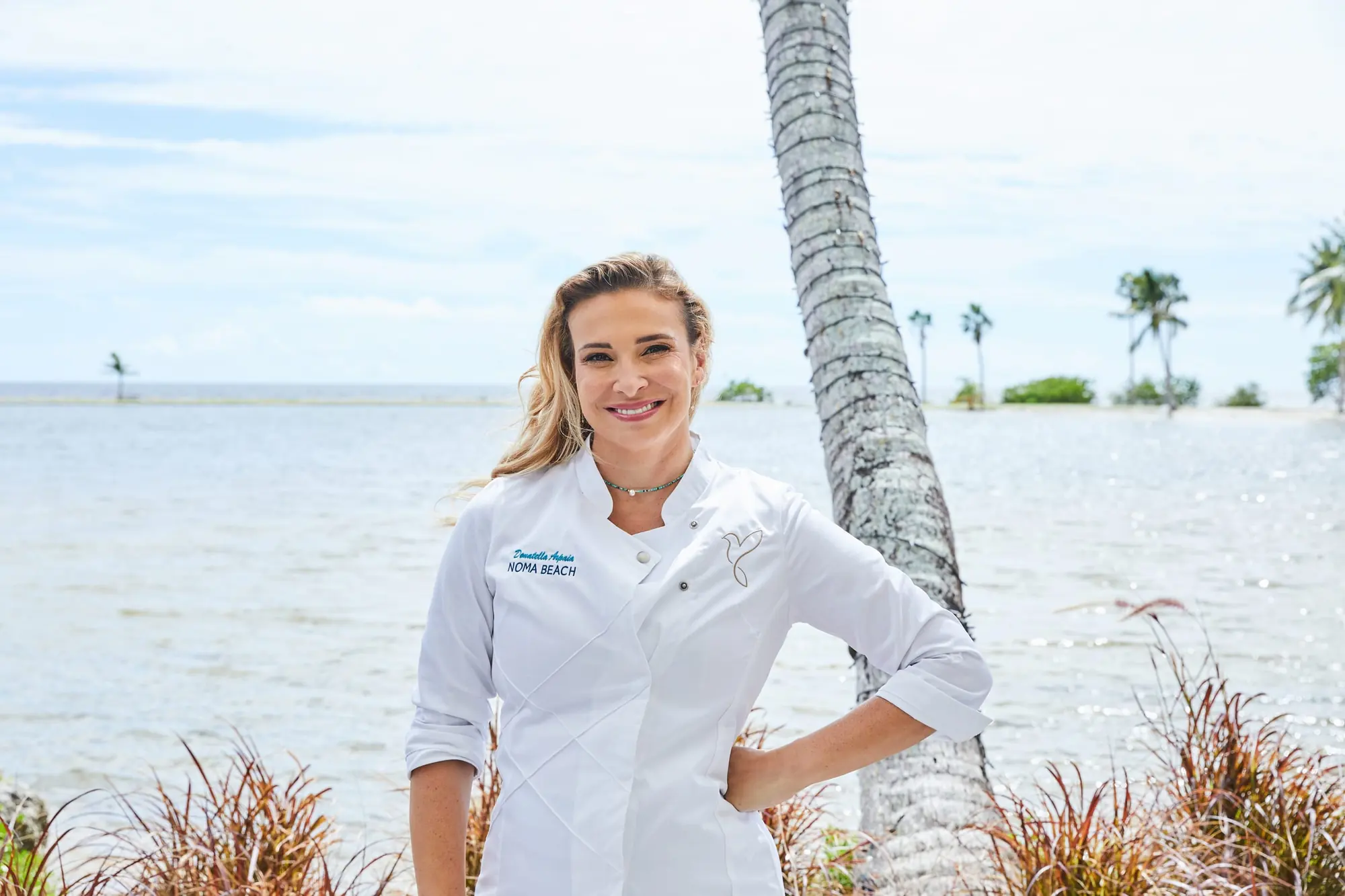 Donatella Arpaia in her NOMA BEACH chef’s jacket by the lagoon, a hand on her hip