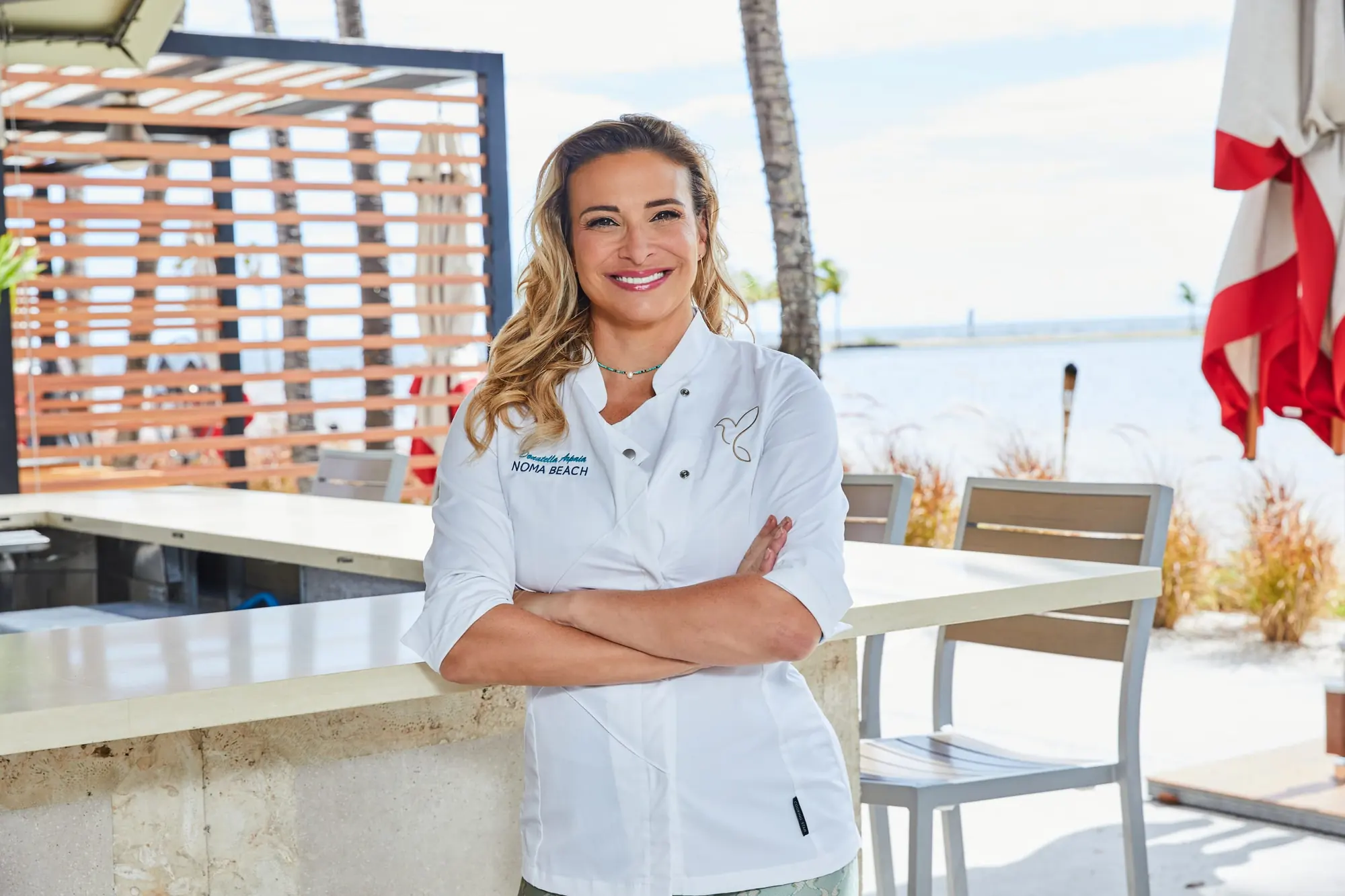 Donatella Arpaia in her NOMA BEACH chef’s jacket, arms crossed and smiling, at the outdoor bar with the lagoon behind her