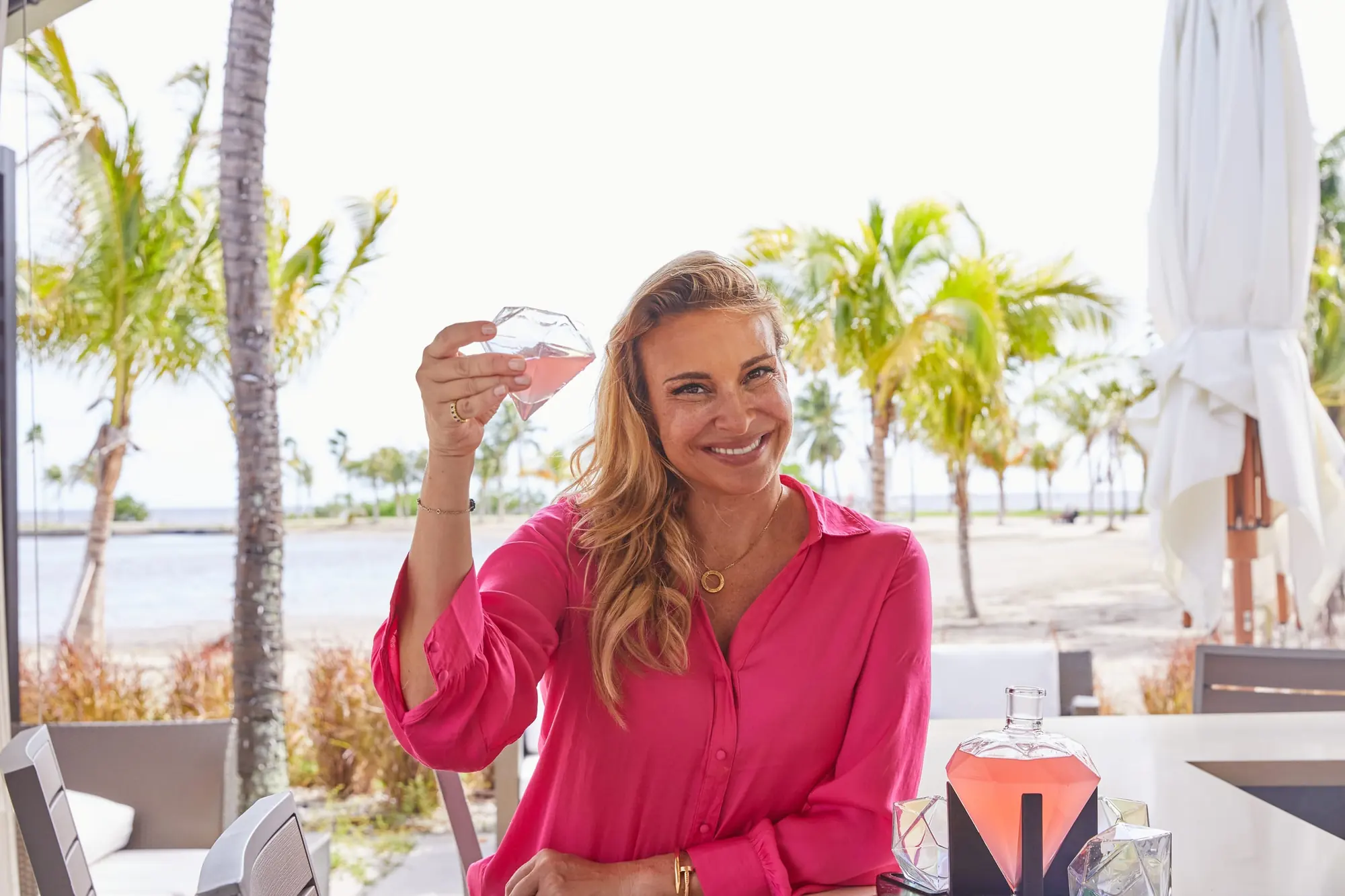 Donatella Arpaia in a pink blouse, raising a glass on the patio, palms and the lagoon behind her