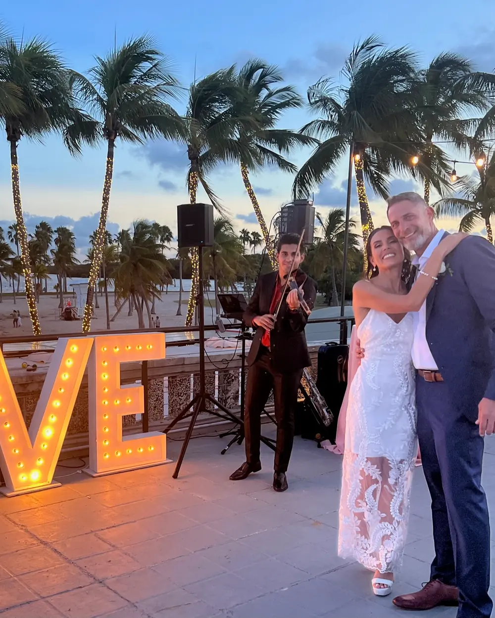 A bride and groom embracing on the terrace beside a lit LOVE sign, a violinist behind them