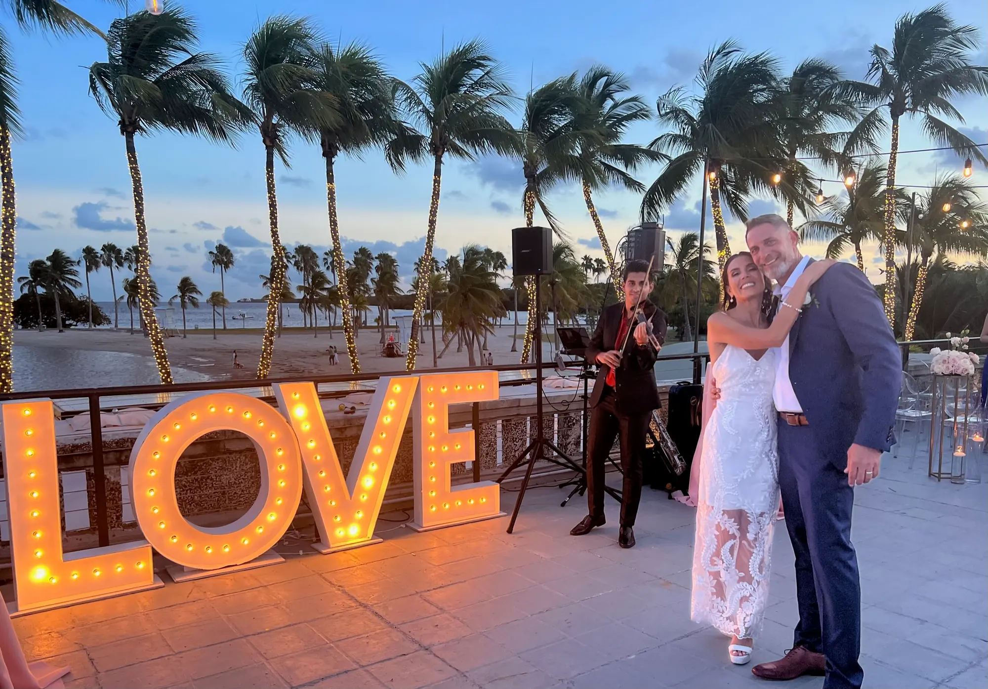 A bride and groom embracing on the terrace beside a lit LOVE sign, a violinist and the palms behind them
