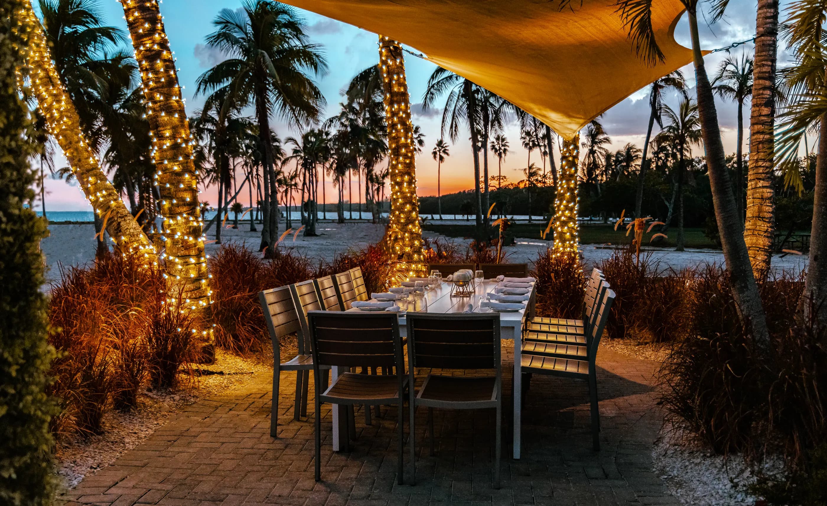 A long table set for dinner under a sail among palms wrapped in lights, the sky over the lagoon turning orange