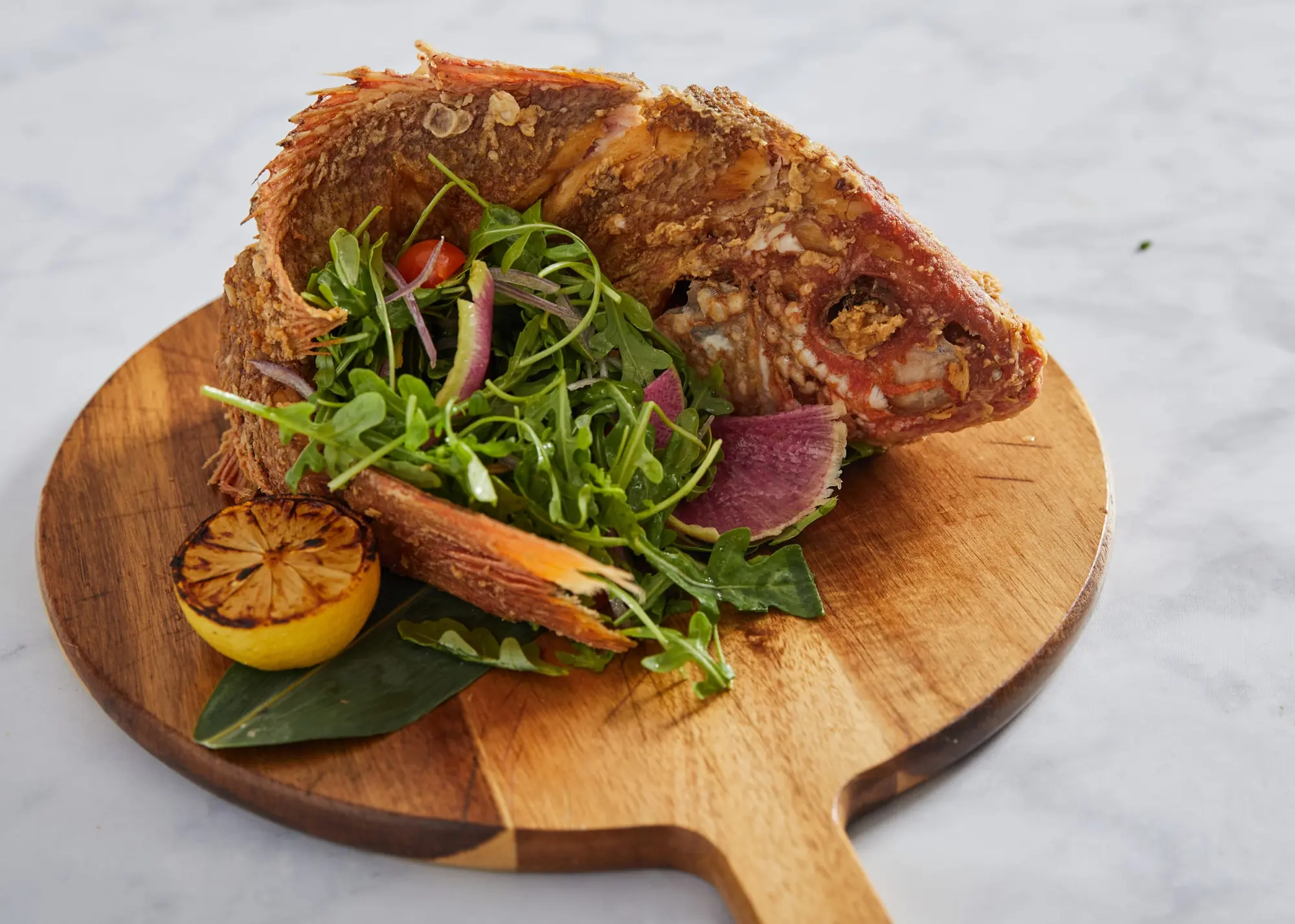 A whole branzino on a wooden board with greens and a grilled lemon