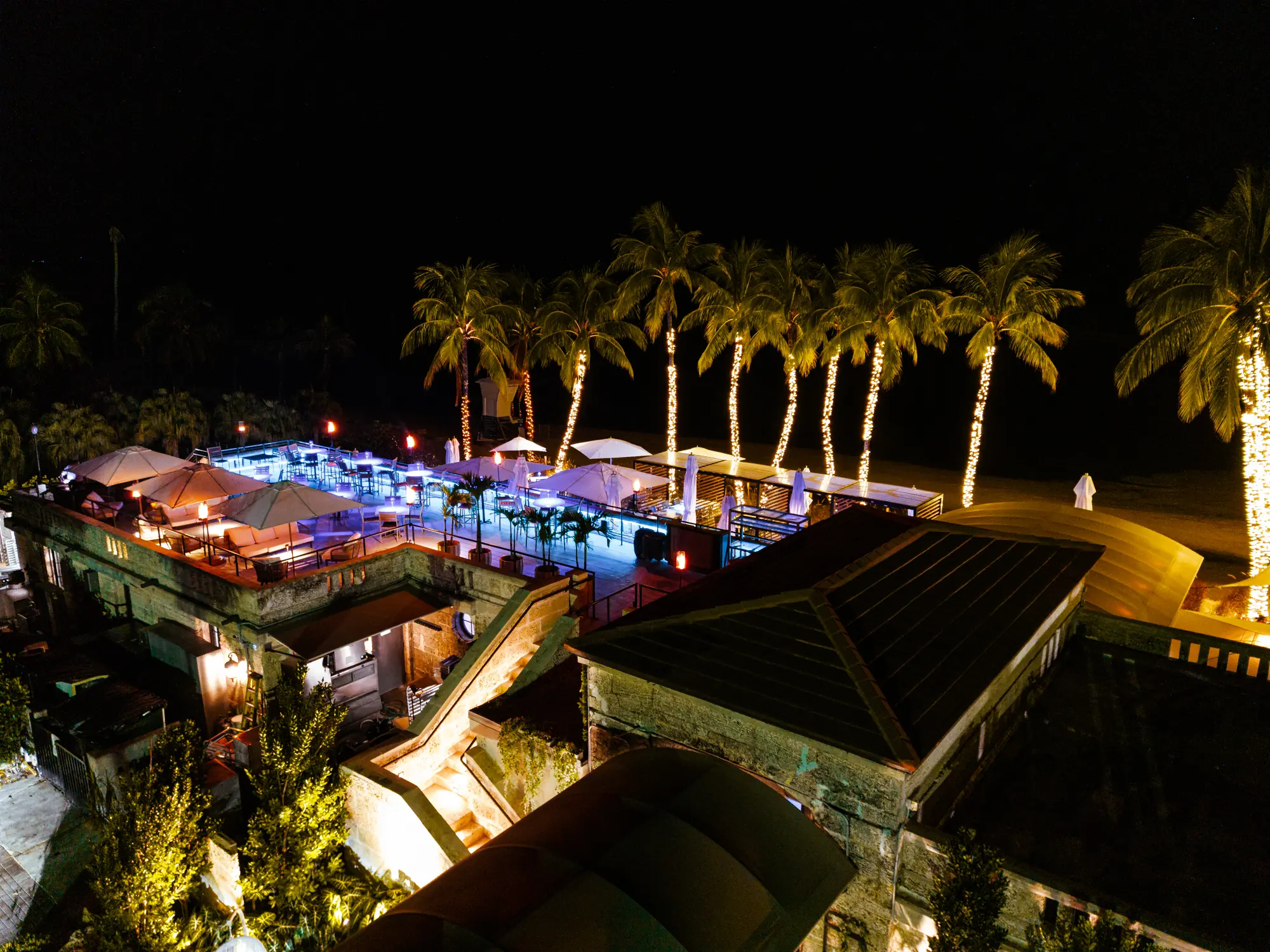 The whole property at night from above, the rooftop lit and the palms in lights