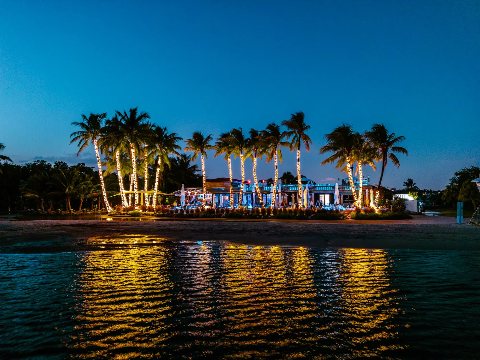 The restaurant at dusk from across the lagoon, its palms in lights and mirrored in the water