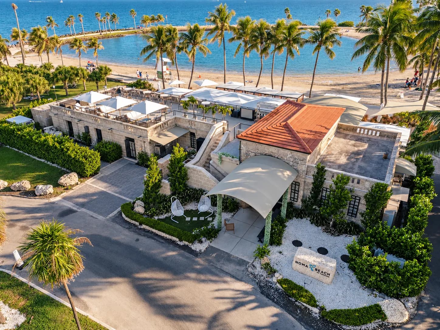 The coral-rock building and its red roof from above, the entrance and its garden, the beach and the bay behind