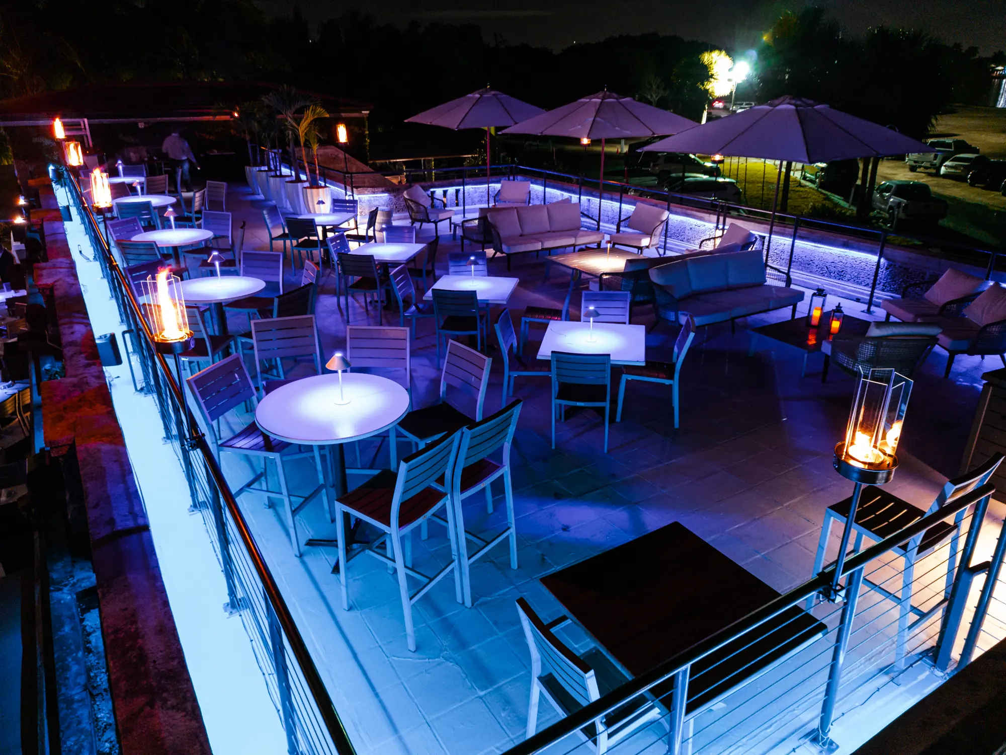 The rooftop at night from above, its tables lit blue and lanterns along the rail
