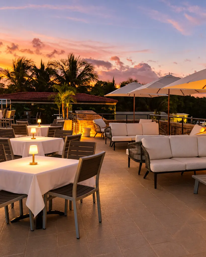 The rooftop terrace at sunset, tables in white linen with little lamps, white sofas under lit umbrellas, pink clouds over the palms