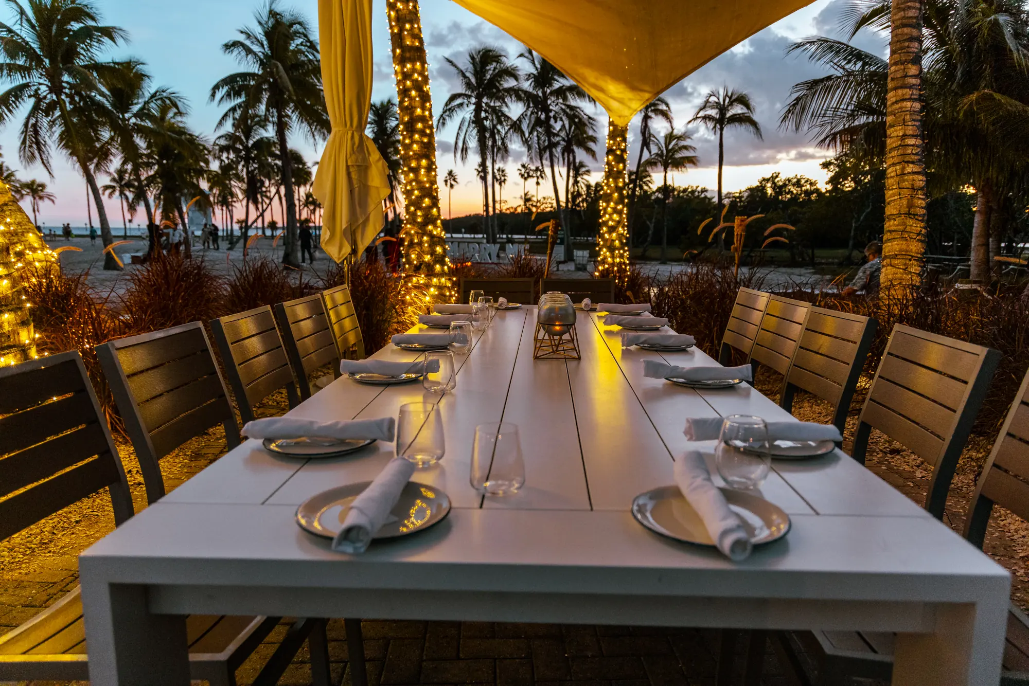 A long table set under a sail at sunset, palms wrapped in lights