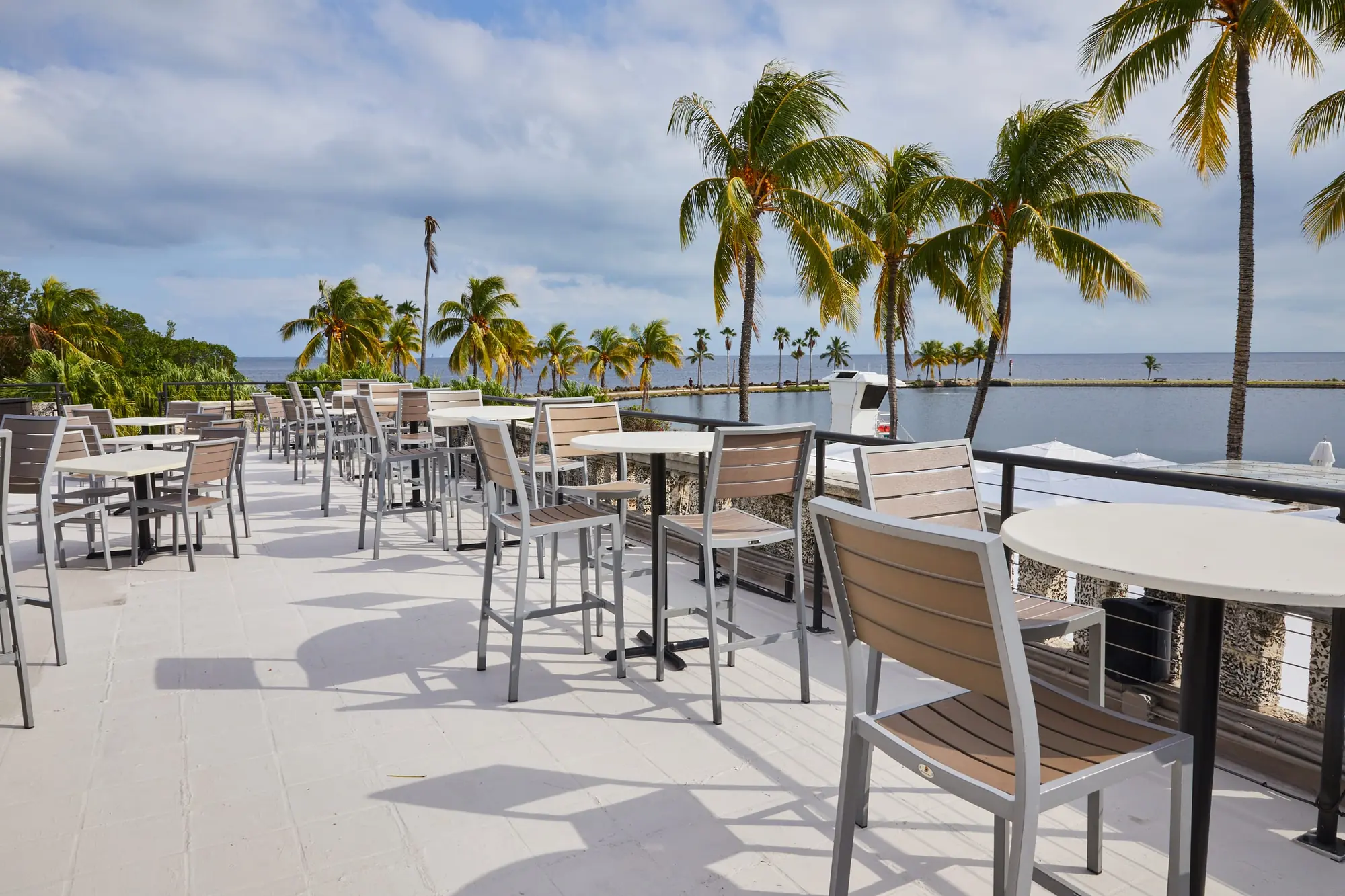 Tables on the terrace by day, the bay and a line of palms beyond the rail