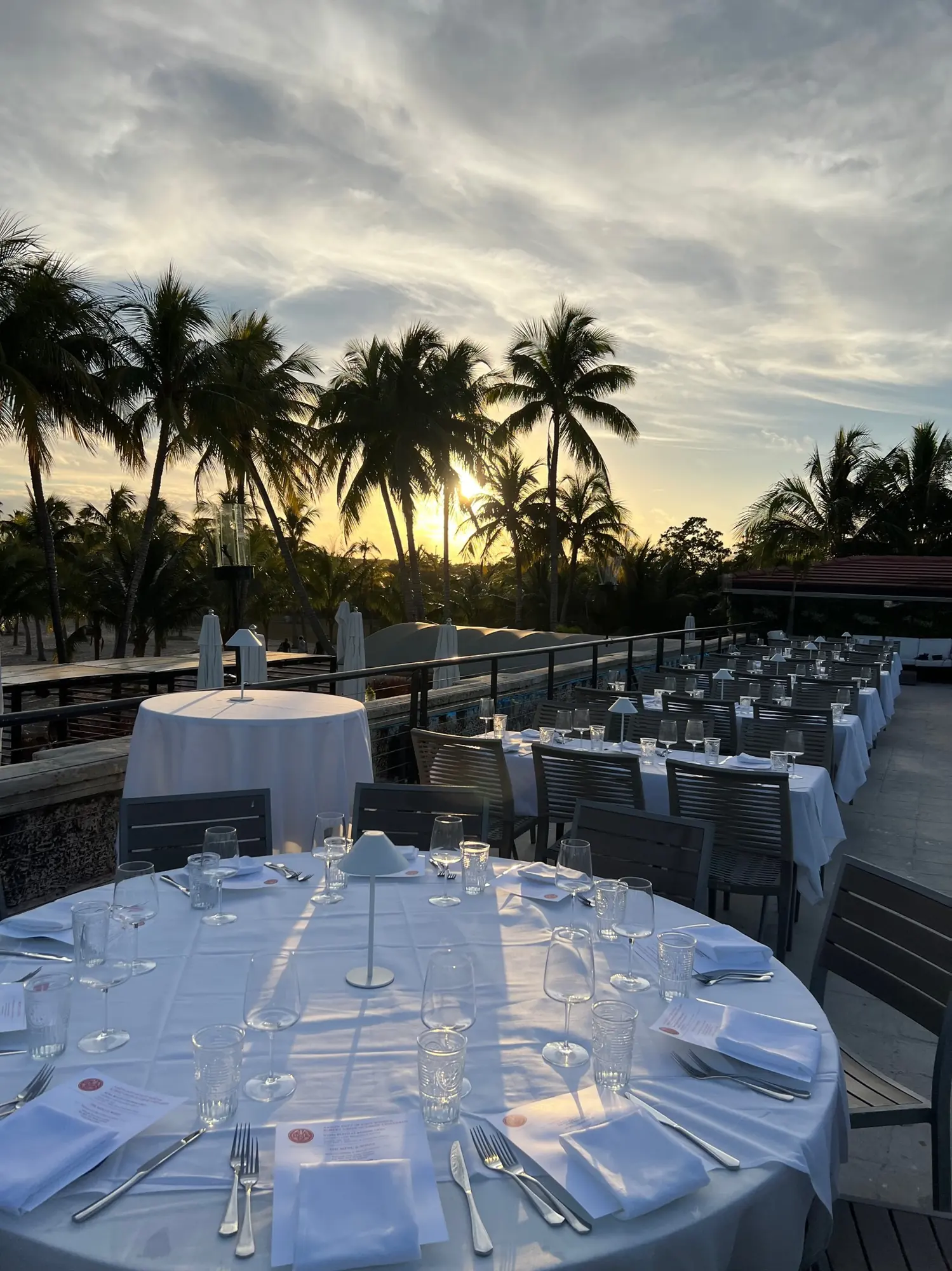 A round table set for dinner on the terrace at dusk, the palms against the evening sky