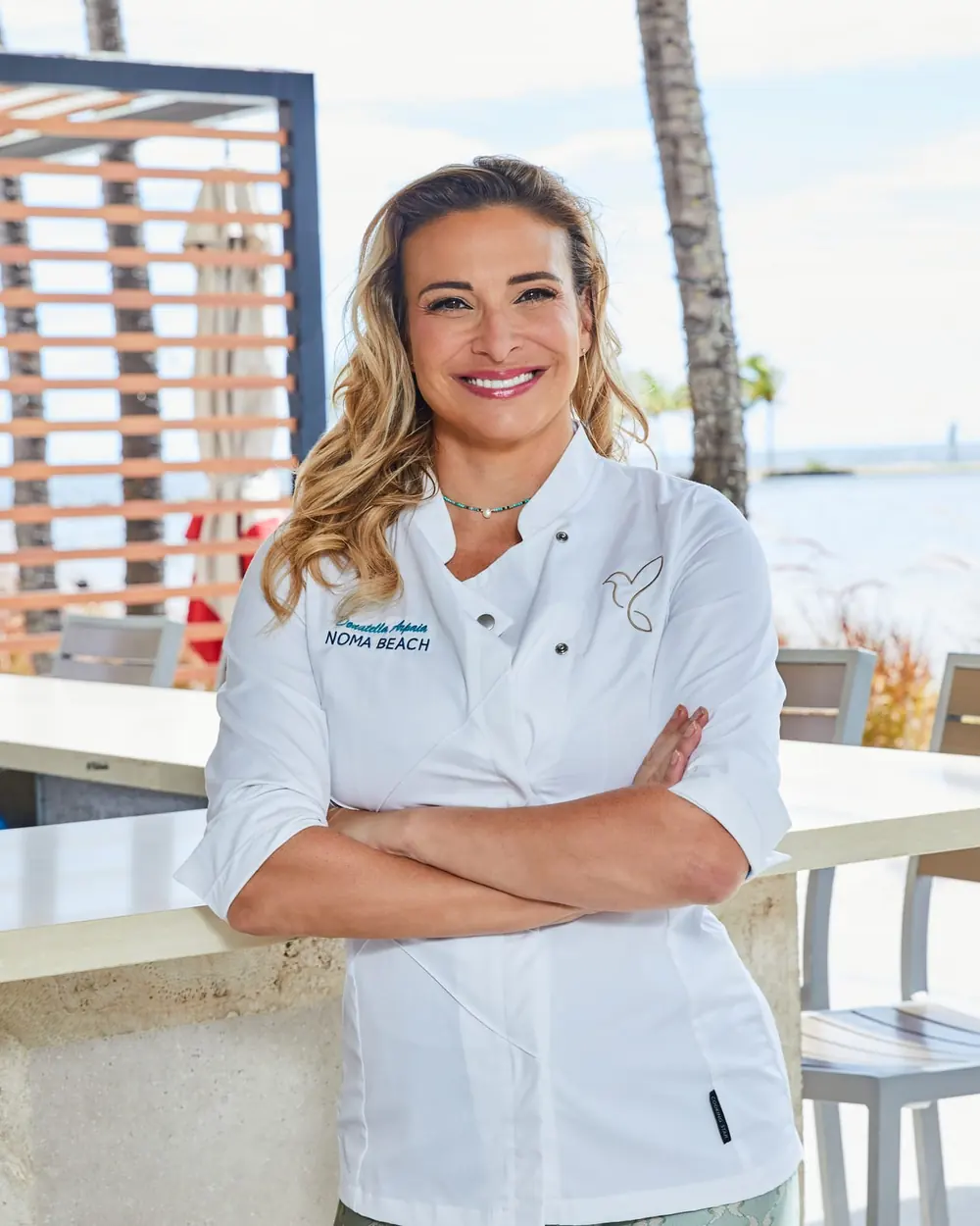 Donatella Arpaia in her NOMA BEACH chef’s jacket, arms crossed and smiling, at the outdoor bar