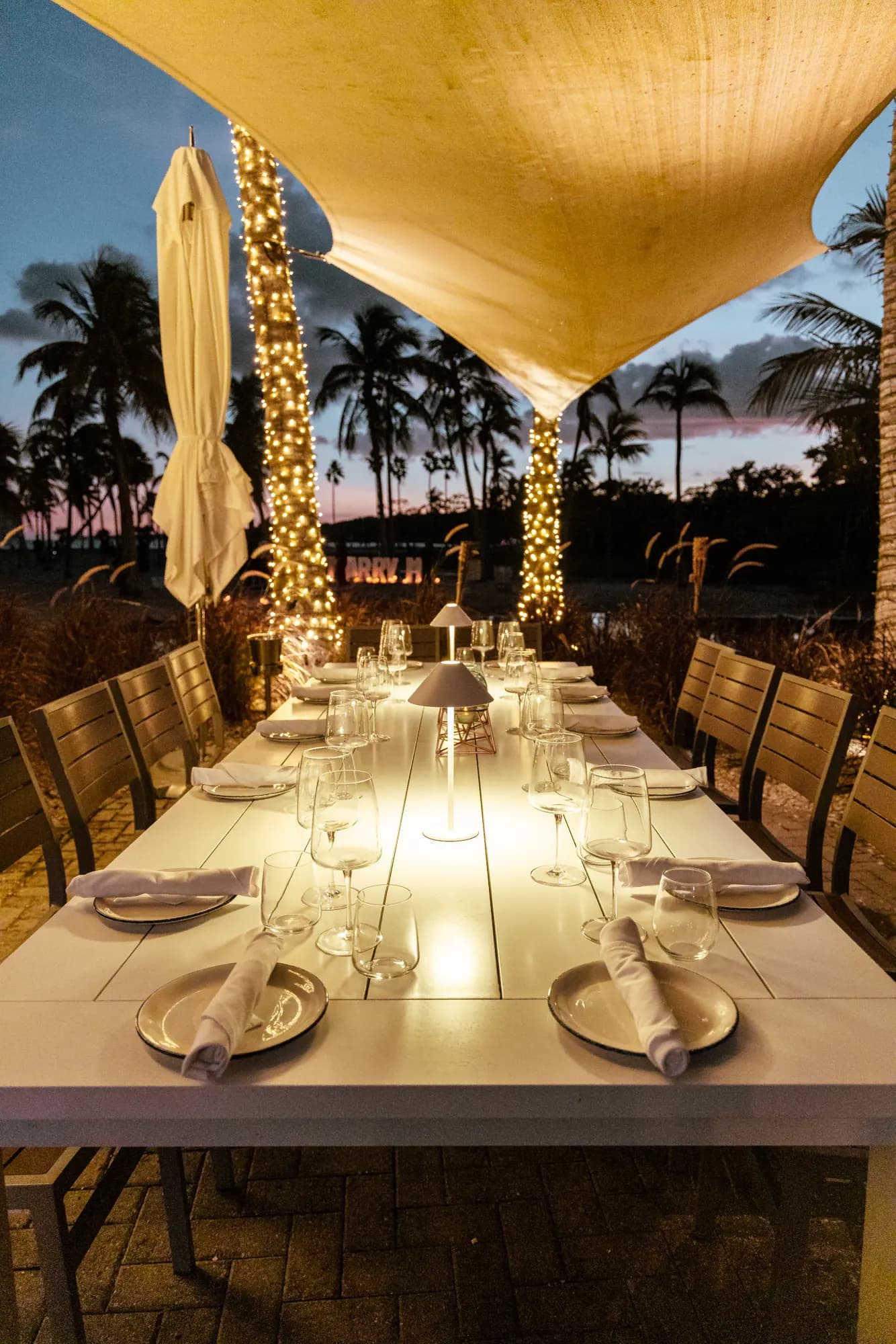 A long table set with glasses and a table lamp, palms in lights against the evening sky